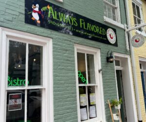 Black sign that reads 'Always Flavored' with painting of black penguin on the left. Green building with two windows with neon signs. Circular sign that reads 'Always Flavored'